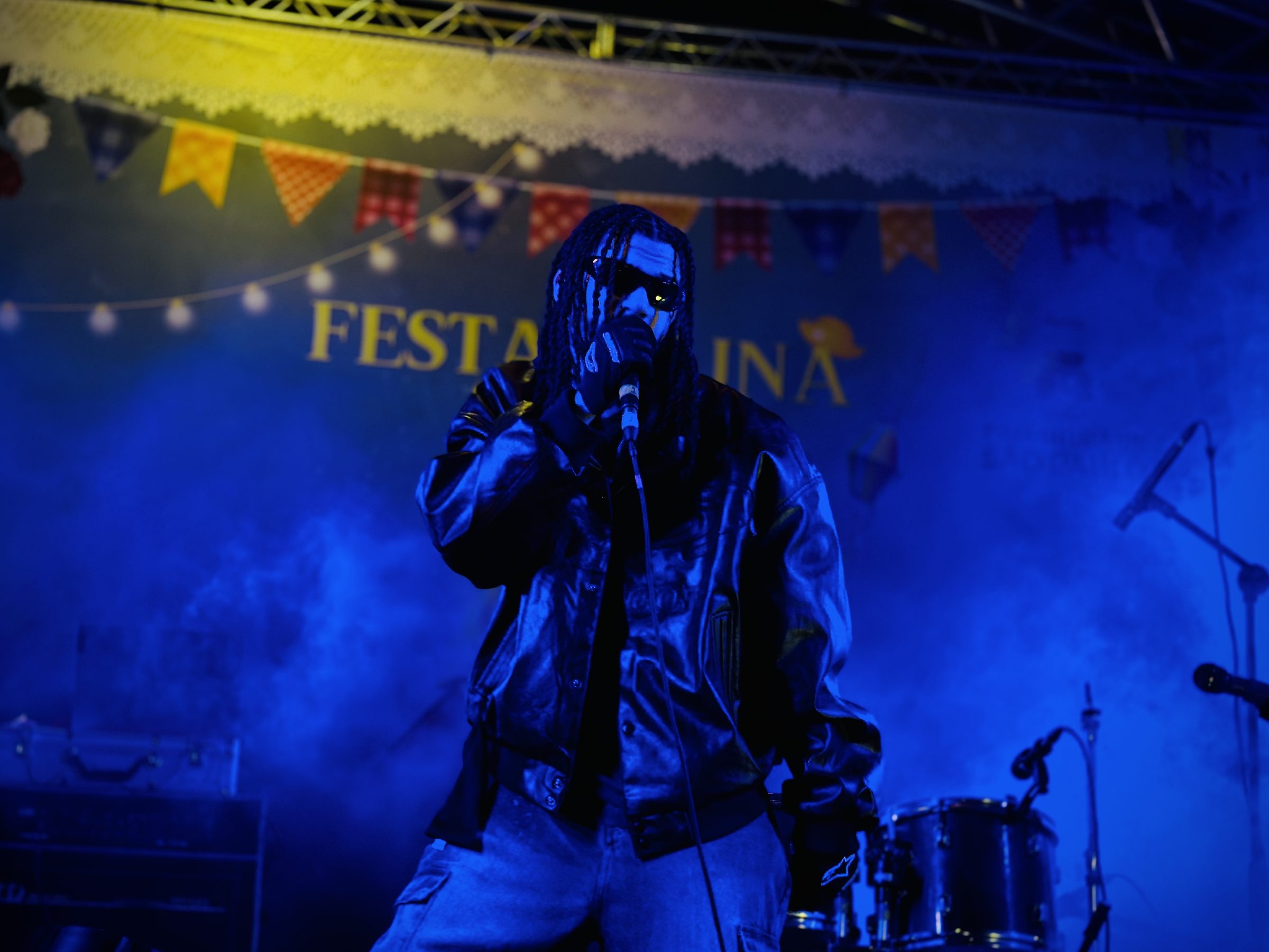BAILON! cantando sob luz azul com bandeirinhas de festa junina ao fundo, 2026